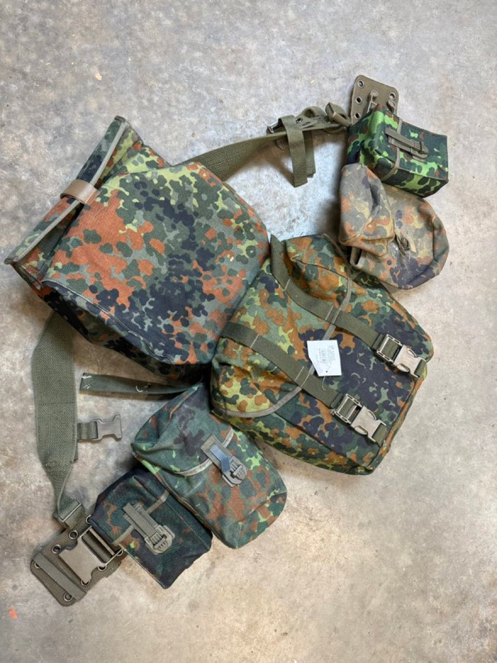 A set of used German military flecktarn patrolling gear including a belt, suspenders, various pouches, a metal canteen, and a gas mask bag.