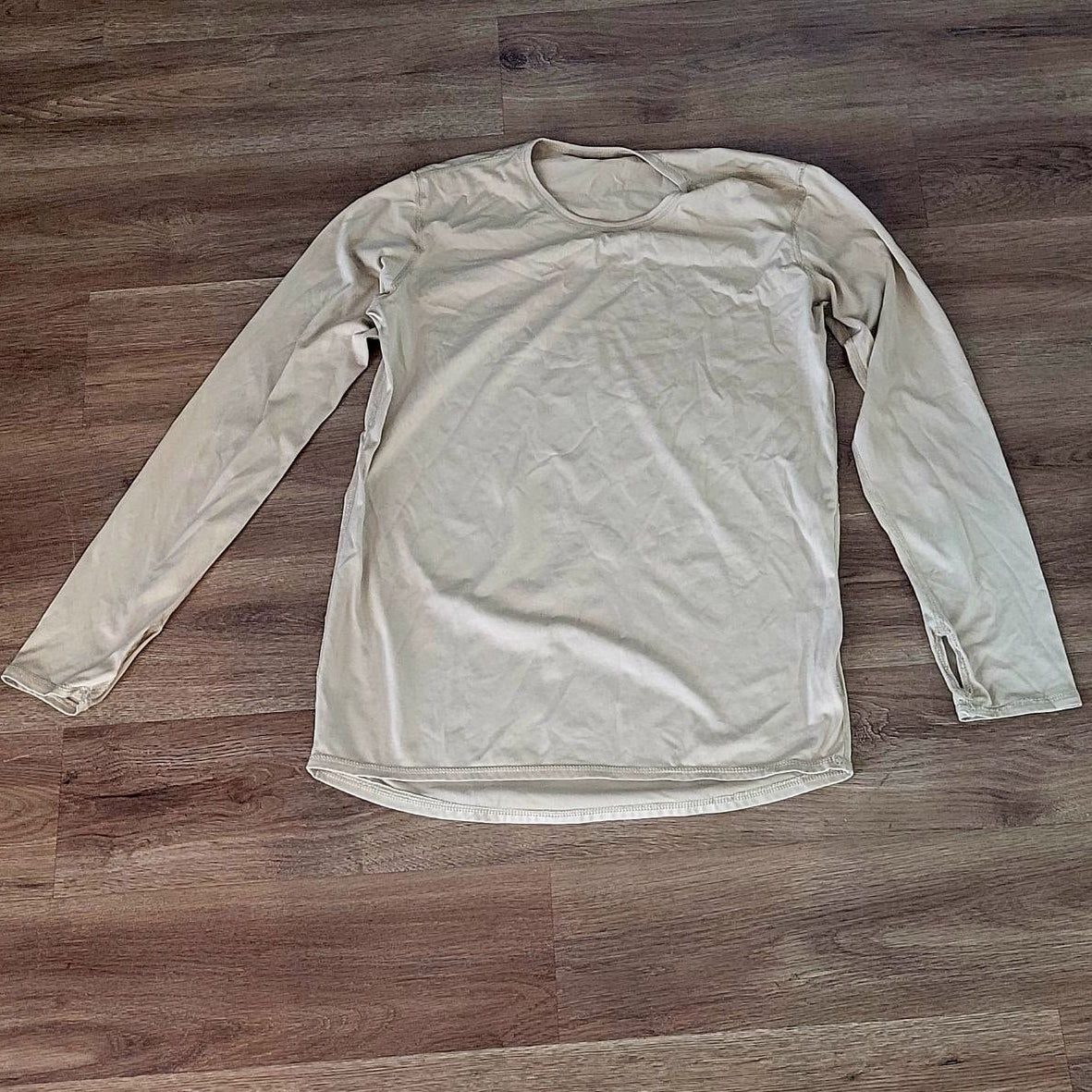 A light beige long-sleeve crewneck shirt laid flat on a brown wood floor.