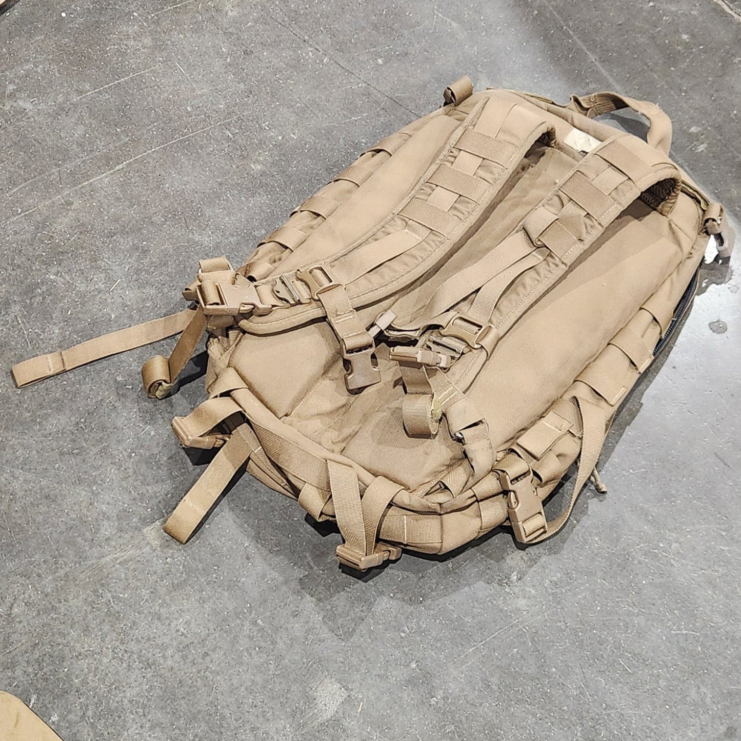 Tan military-style backpack on a concrete floor with shoes nearby