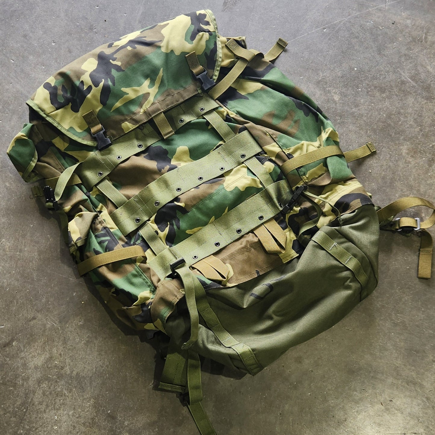 Camouflage backpack on a concrete floor with a person's foot visible.