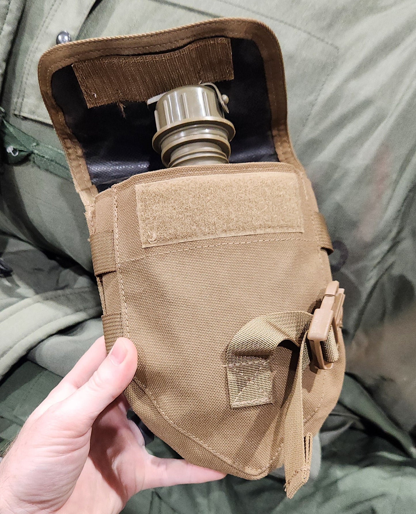 Person holding a tactical gas mask pouch with a gas mask inside, on a green military-style background.