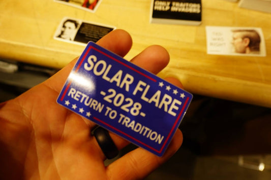 Hand holding a blue and red sticker with text 'Solar Flare 2028 Return to Tradition' against a blurred background.