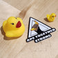 Two small yellow rubber ducks on a wooden surface with a black and white triangle-shaped object featuring text.