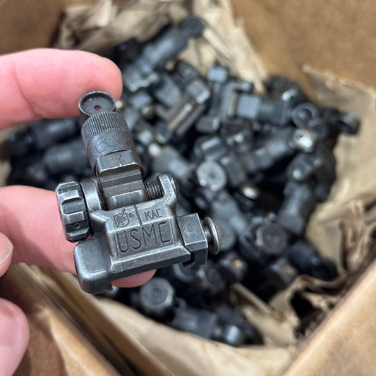 Hand holding a metal valve handle with 'USMC' branding, in front of a cardboard box filled with similar valves.