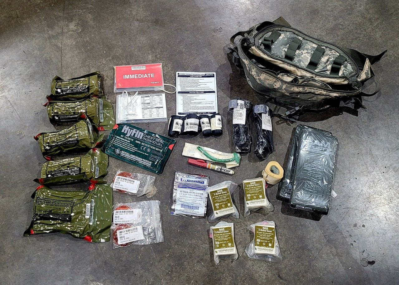 A tactical medical survival kit laid out on a concrete floor, featuring a camouflage backpack, multiple olive green hydration packs, medical gauze, bandages, a green Hytm cooling pack, and other emergency supplies.