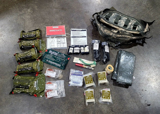 A tactical medical survival kit laid out on a concrete floor, featuring a camouflage backpack, multiple olive green hydration packs, medical gauze, bandages, a green Hytm cooling pack, and other emergency supplies.