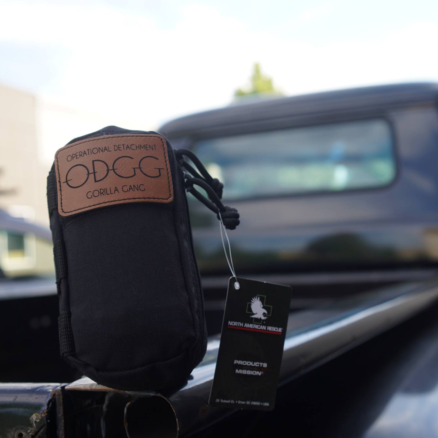 A black ODGG Gorilla Gant pouch with a brown leather patch, resting on the tailgate of a black pickup truck. A black hangtag is attached to the pouch.