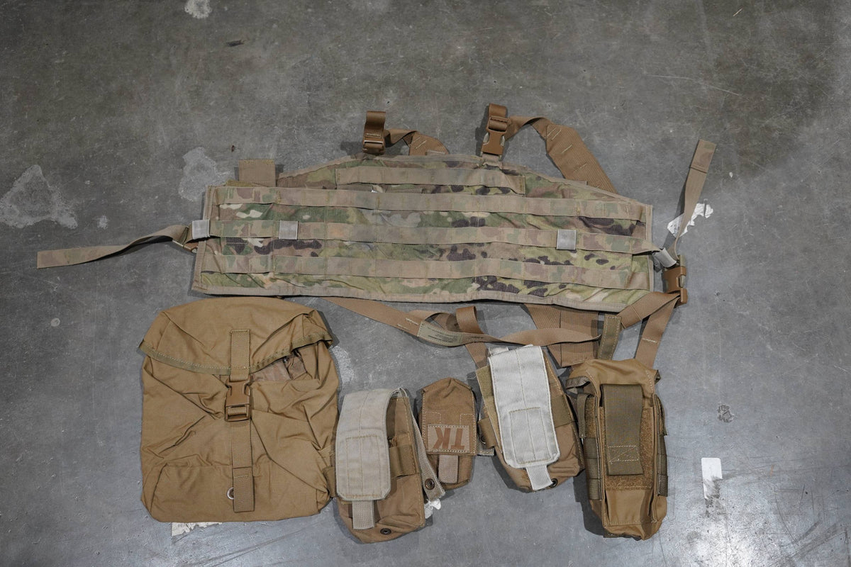 A tactical gear set laid out on a gray concrete floor, including a camouflage MOLLE webbing belt, a tan backpack, and several matching pouches and straps.
