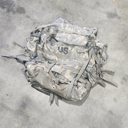 A camouflage military-style backpack or gear bag lying on a concrete floor, with straps and buckles visible. Parts of a person's legs and shoes are visible at the edges of the frame.