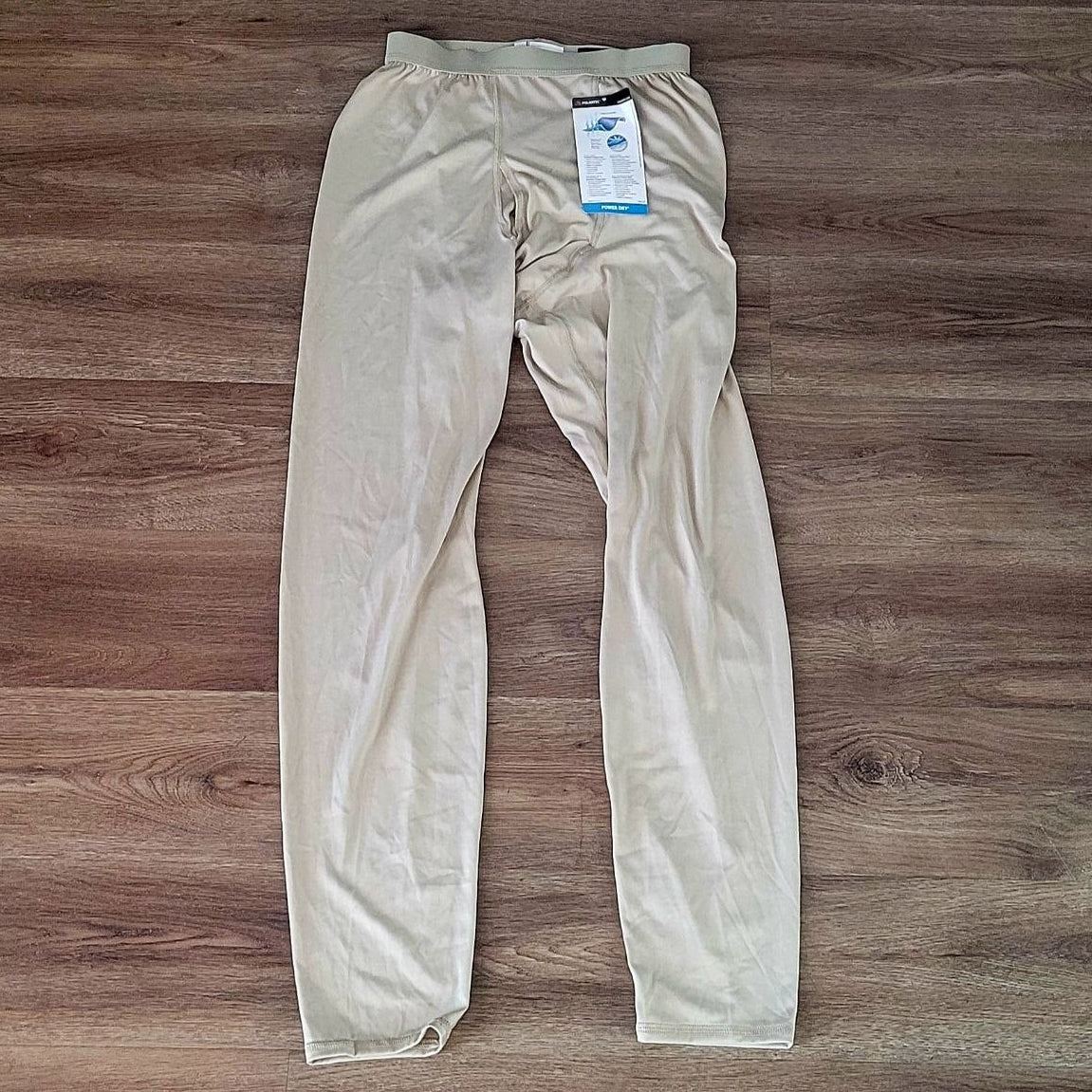 A pair of light beige compression pants laid flat on a brown wood-look floor, with a product tag still attached near the waistband.