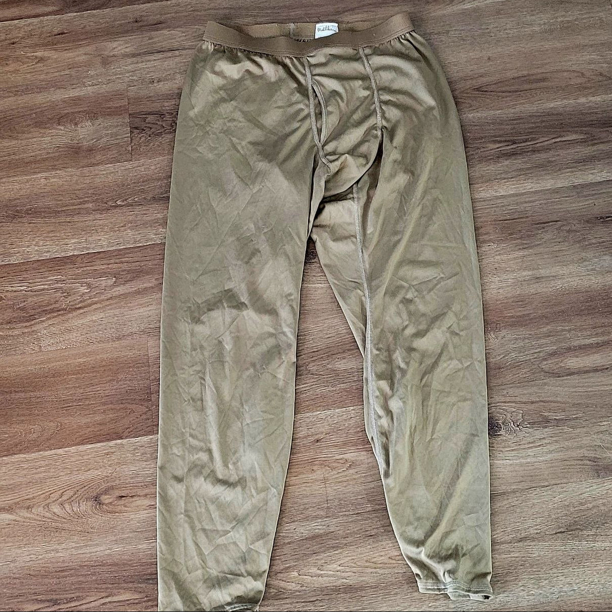 A pair of loose-fitting, lightweight pants in a muted khaki-beige color, laid flat on a brown wood-look floor.