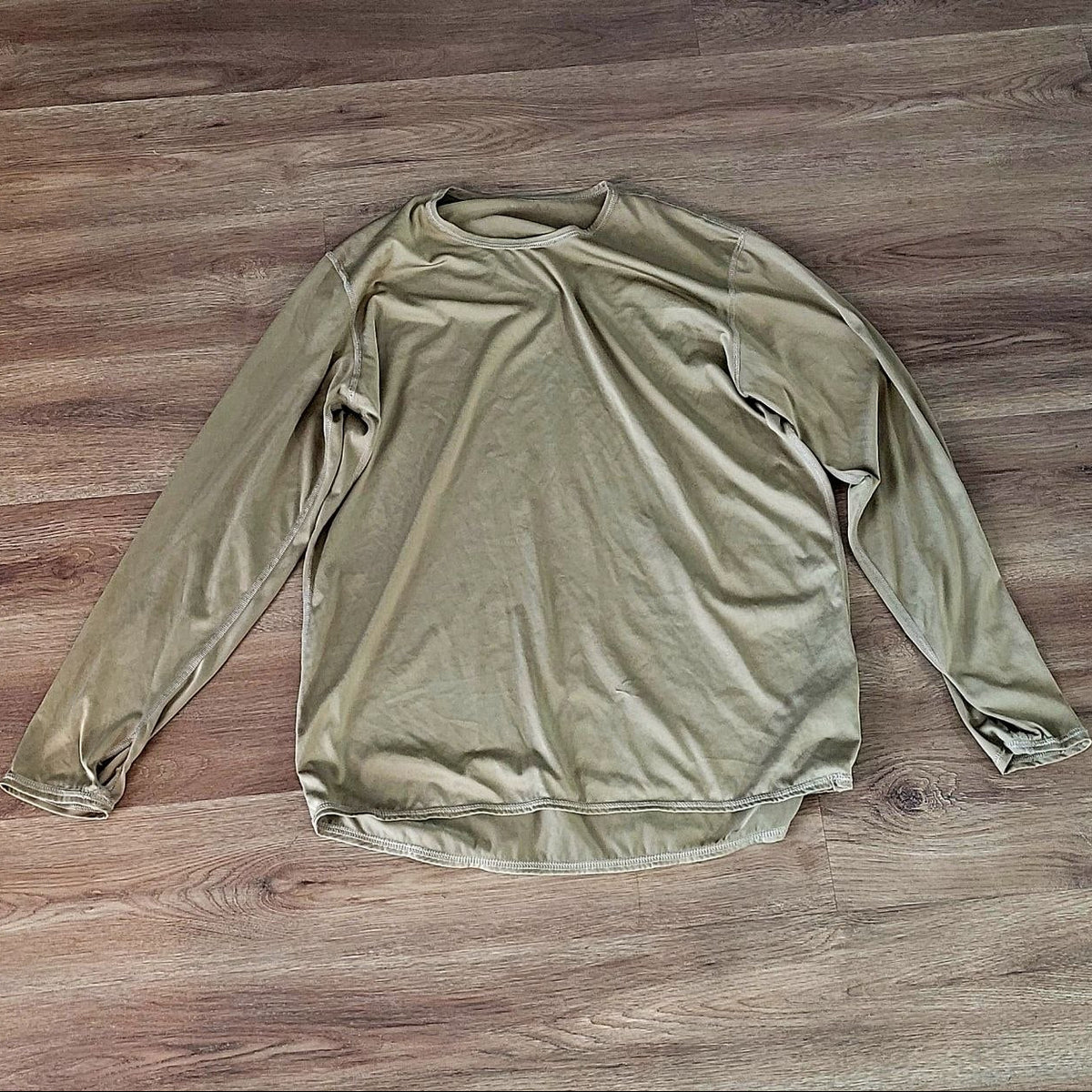 A long-sleeve crewneck shirt in a muted olive green color, laid flat on a brown wood plank floor.