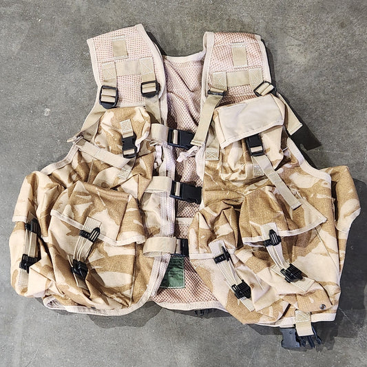 Camouflage tactical vest on a concrete floor with a person's feet visible.