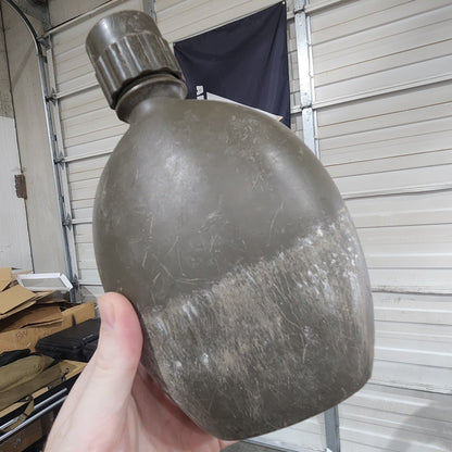 A person's hand holding a dirty, worn military-style canteen bottle in a garage setting with a white roll-up door in the background.