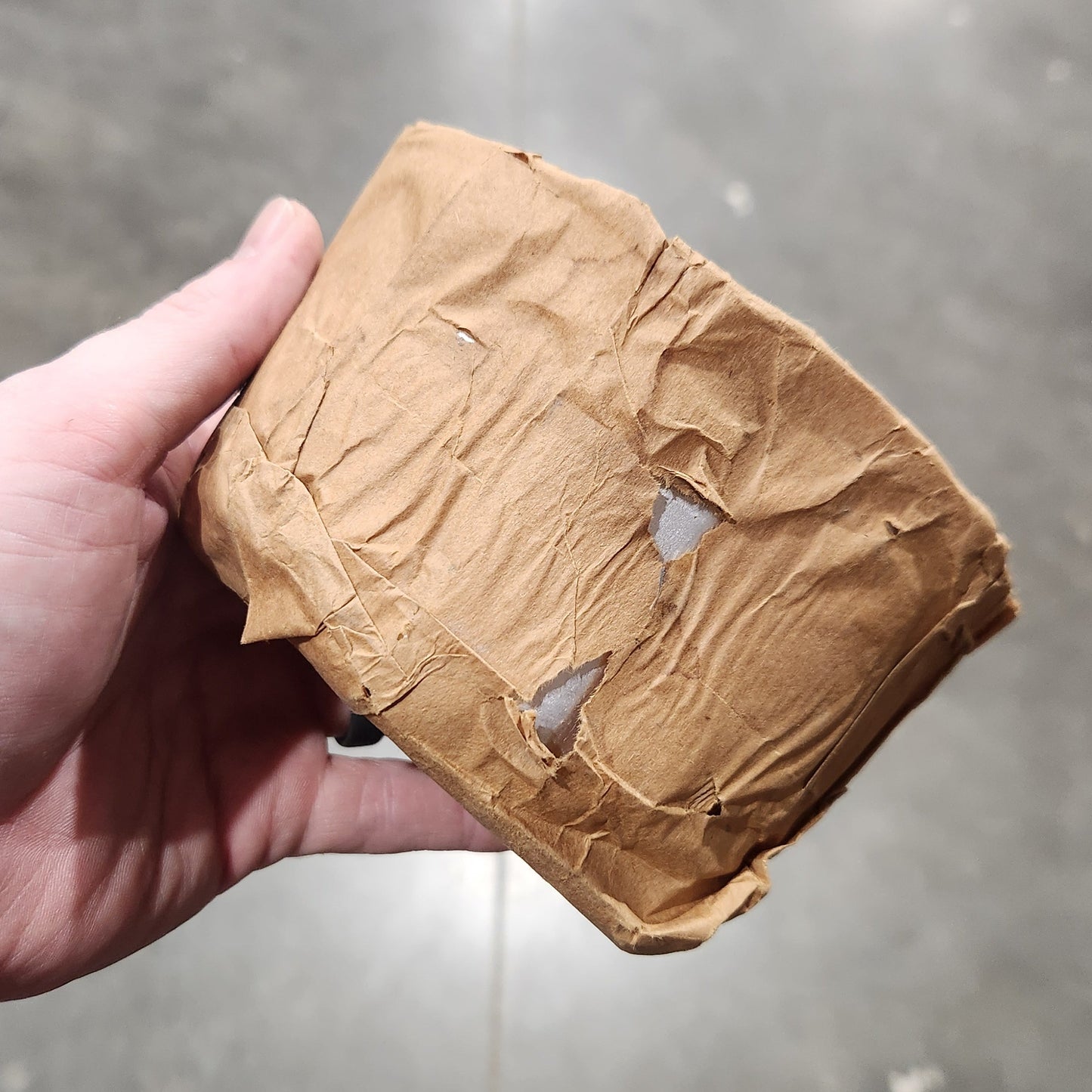 A hand holding a crumpled brown paper bag against a gray concrete floor background.