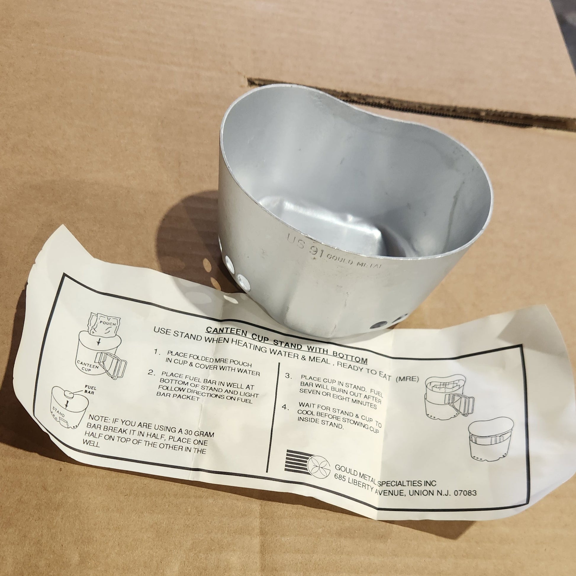 A silver metal elbow pipe fitting placed next to its white paper instruction sheet on a brown cardboard surface.