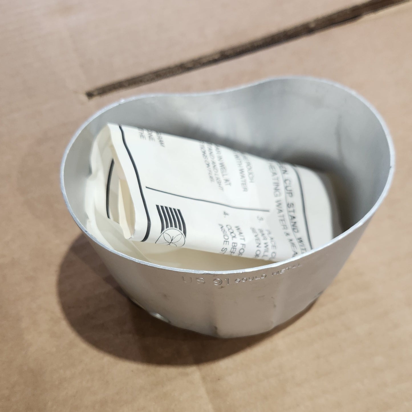 A silver metal cup stand with a white paper instruction sheet inside, resting on a brown cardboard box.
