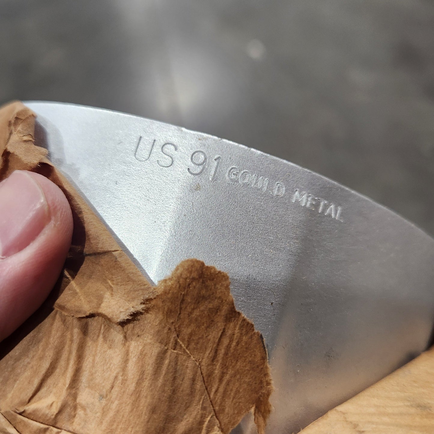 A close-up of a curved silver metal knife blade with 'US 91 GOULD METAL' engraved on it, held by a hand against a blurred gray background with brown paper wrapping visible on the right.
