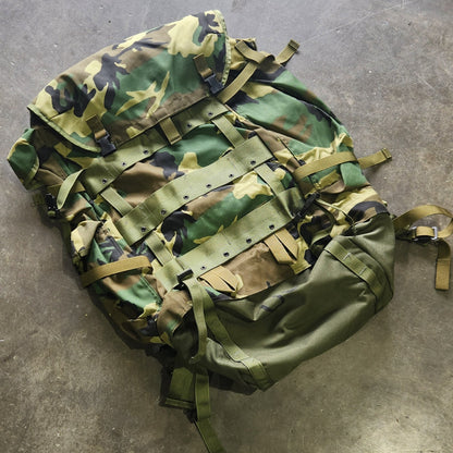 Camouflage backpack on a concrete floor with a person's foot visible.
