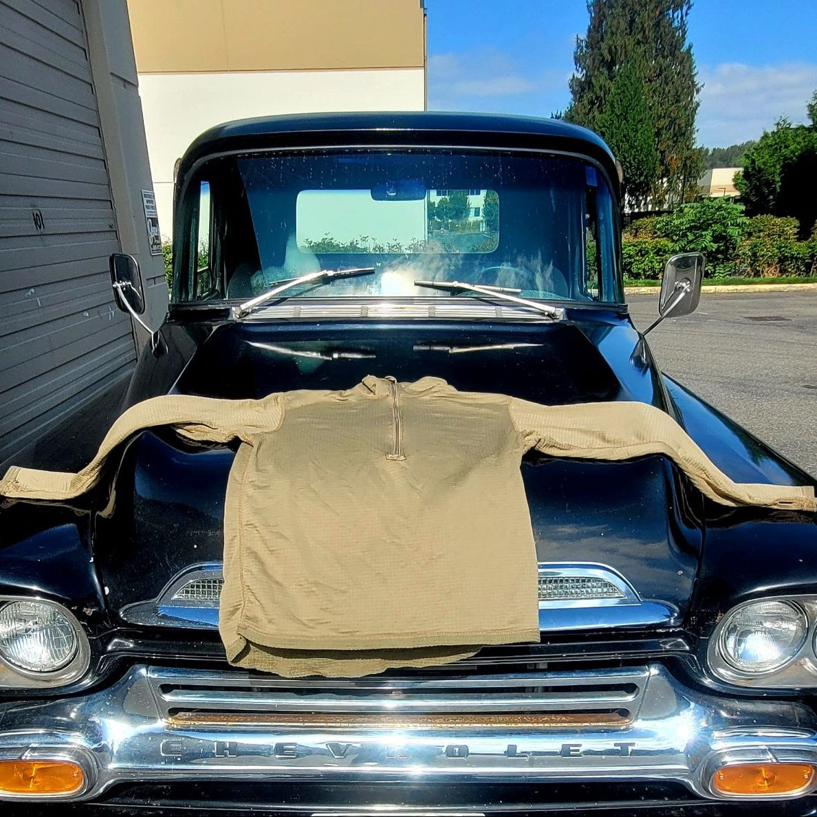 A tan long-sleeve half-zip pullover shirt is draped across the front of a black vintage Chevrolet truck outdoors.