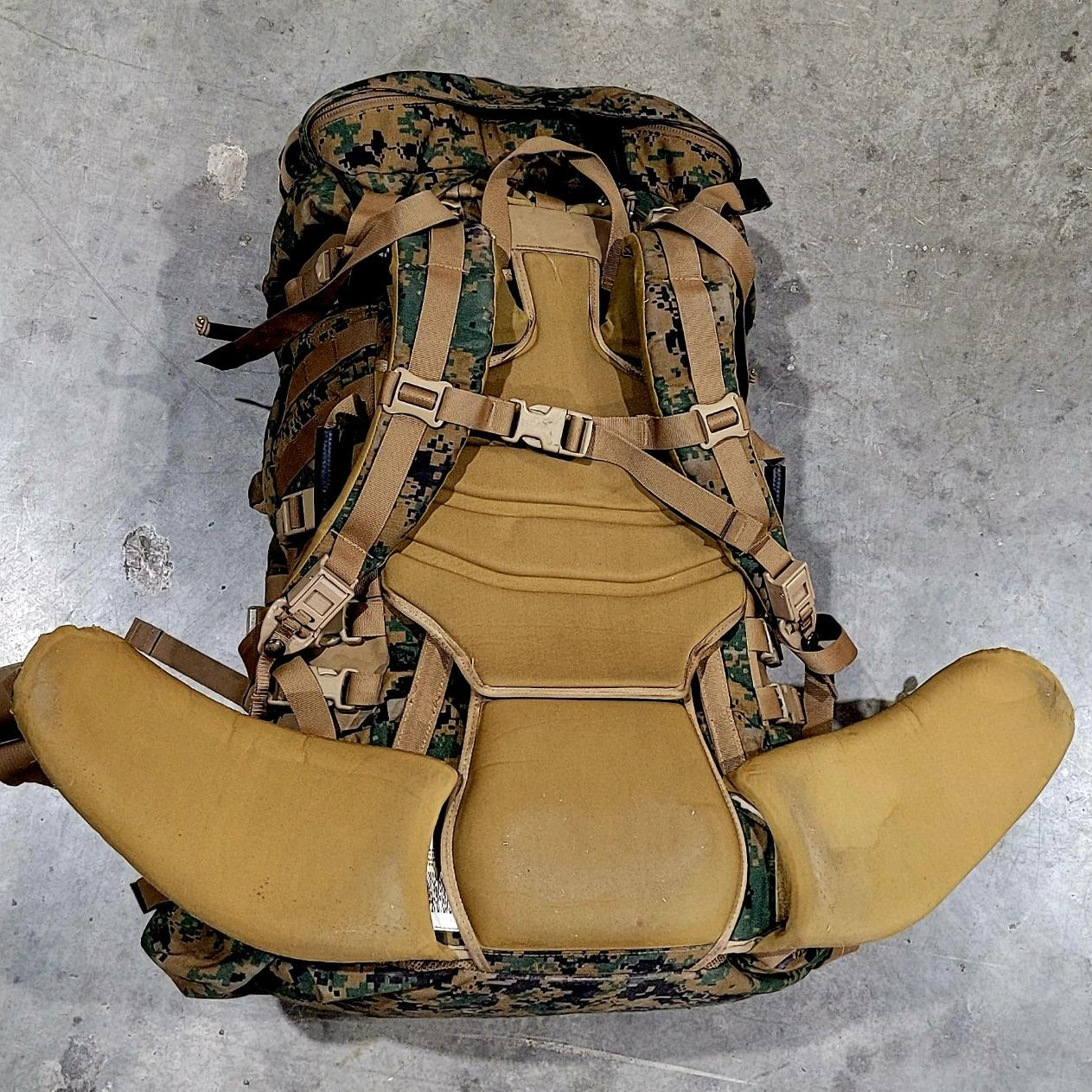 Camouflage backpack with a tan seat pad on a concrete floor
