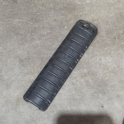 Several black cylindrical grip or handle components arranged on a gray concrete floor, with a cardboard box edge visible on the left and a shadow on the right.