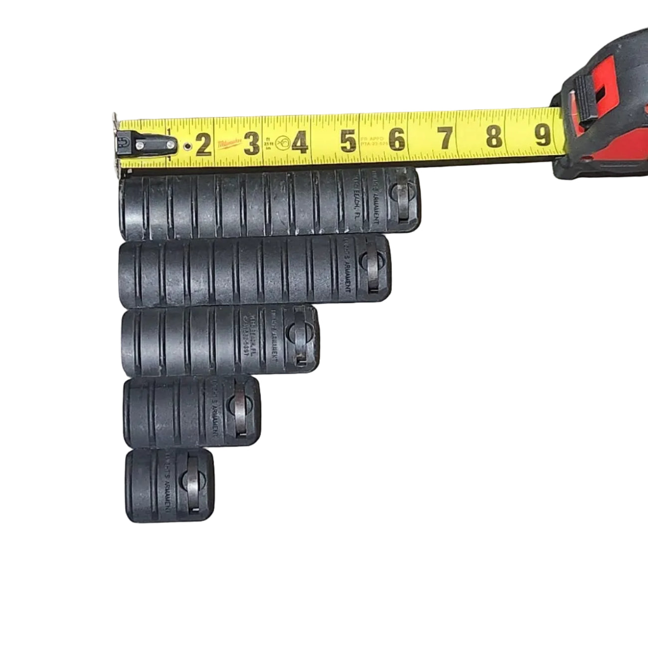 Several black cylindrical plastic pieces arranged on a concrete floor next to a yellow tape measure, with a cardboard box edge visible on the left and a shoe shadow on the right.