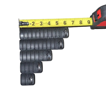 Several black cylindrical plastic pieces arranged on a concrete floor next to a yellow tape measure, with a cardboard box edge visible on the left and a shoe shadow on the right.