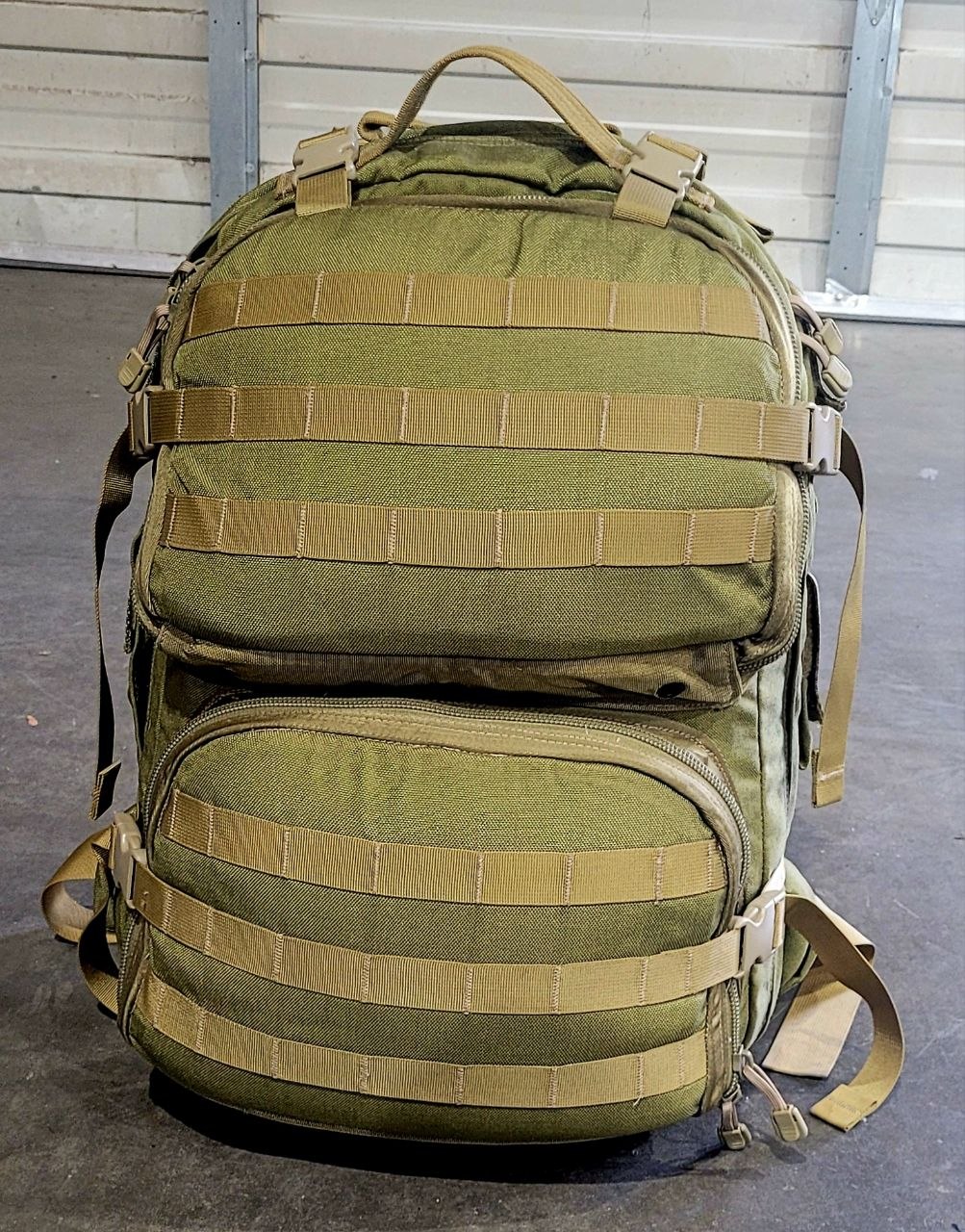Green tactical backpack with multiple straps on a gray floor.