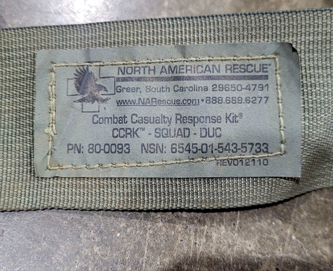 Close-up of a sewn fabric label reading NORTH AMERICAN RESCUE with contact information and Combat Casualty Response Kit text, attached to olive drab fabric.