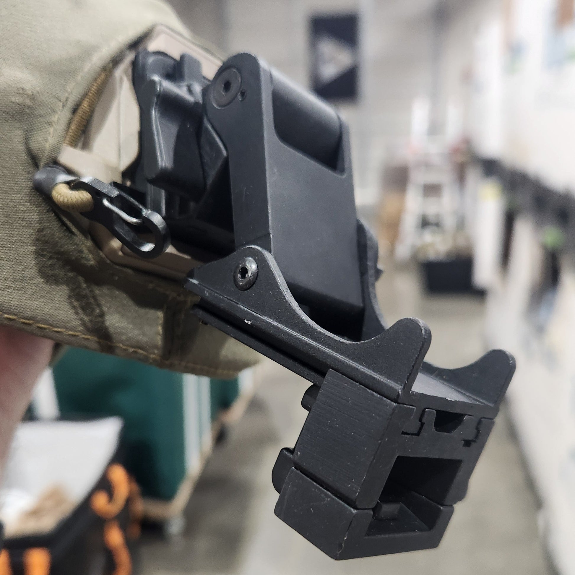 A close-up of a black plastic tactical clip or attachment mounted on a tan fabric strap, held in a hand against a blurred indoor background.