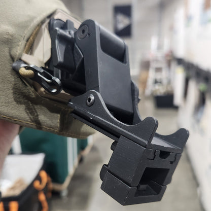 A close-up of a black plastic tactical clip or attachment mounted on a tan fabric strap, held in a hand against a blurred indoor background.