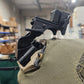 A close-up of a black tactical mounting bracket attached to a tan canvas pouch, photographed in a real indoor warehouse or storage environment with blurred lights and shelving in the background.