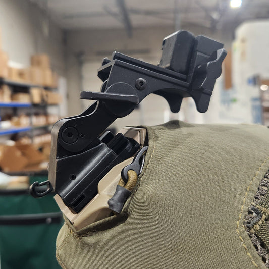 A close-up of a black tactical mounting bracket attached to a tan canvas pouch, photographed in a real indoor warehouse or storage environment with blurred lights and shelving in the background.