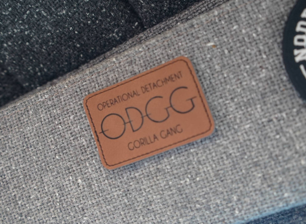 Close-up of a gray woven fabric product with a tan leather patch reading 'OPERATIONAL DETACHMENT ODGG CORILLA CANG', alongside dark textured material and part of a black circular patch.