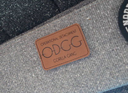Close-up of a gray woven fabric product with a tan leather patch reading 'OPERATIONAL DETACHMENT ODGG CORILLA CANG', alongside dark textured material and part of a black circular patch.