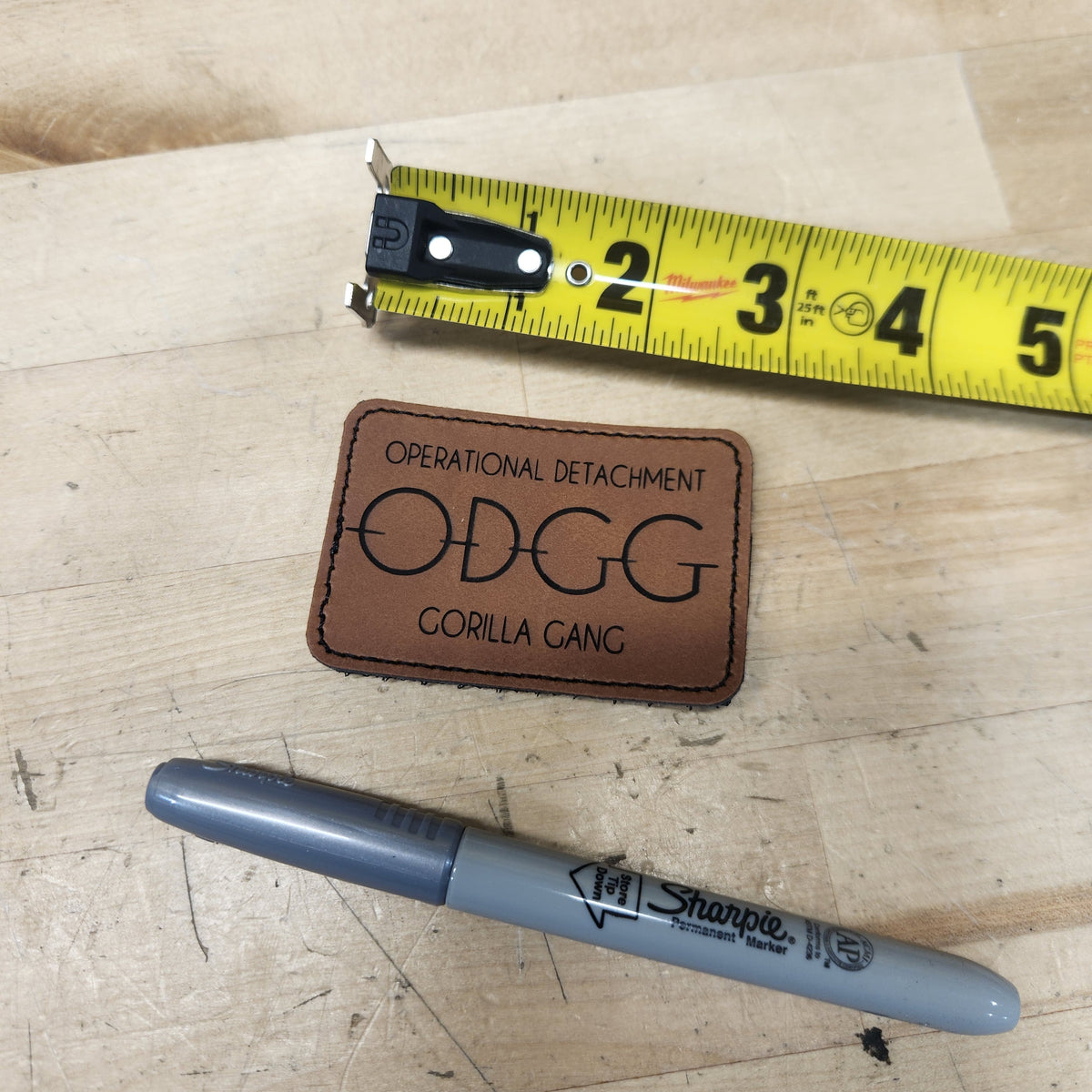 A brown faux-leather patch with 'OPERATIONAL DETACHMENT ODGG CORILLA GANG' printed in black, shown on a light wood surface next to a yellow tape measure and a gray Sharpie marker.