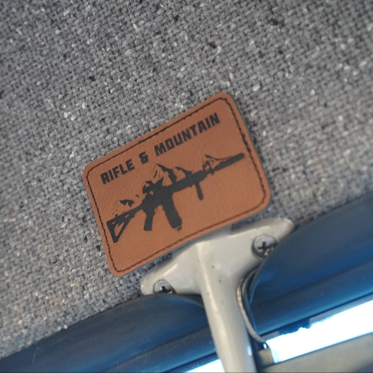A close-up shot of a rectangular brown leather patch with a black rifle silhouette and the text 'RIFLE & MOUNTAIN' on a gray textured fabric surface.
