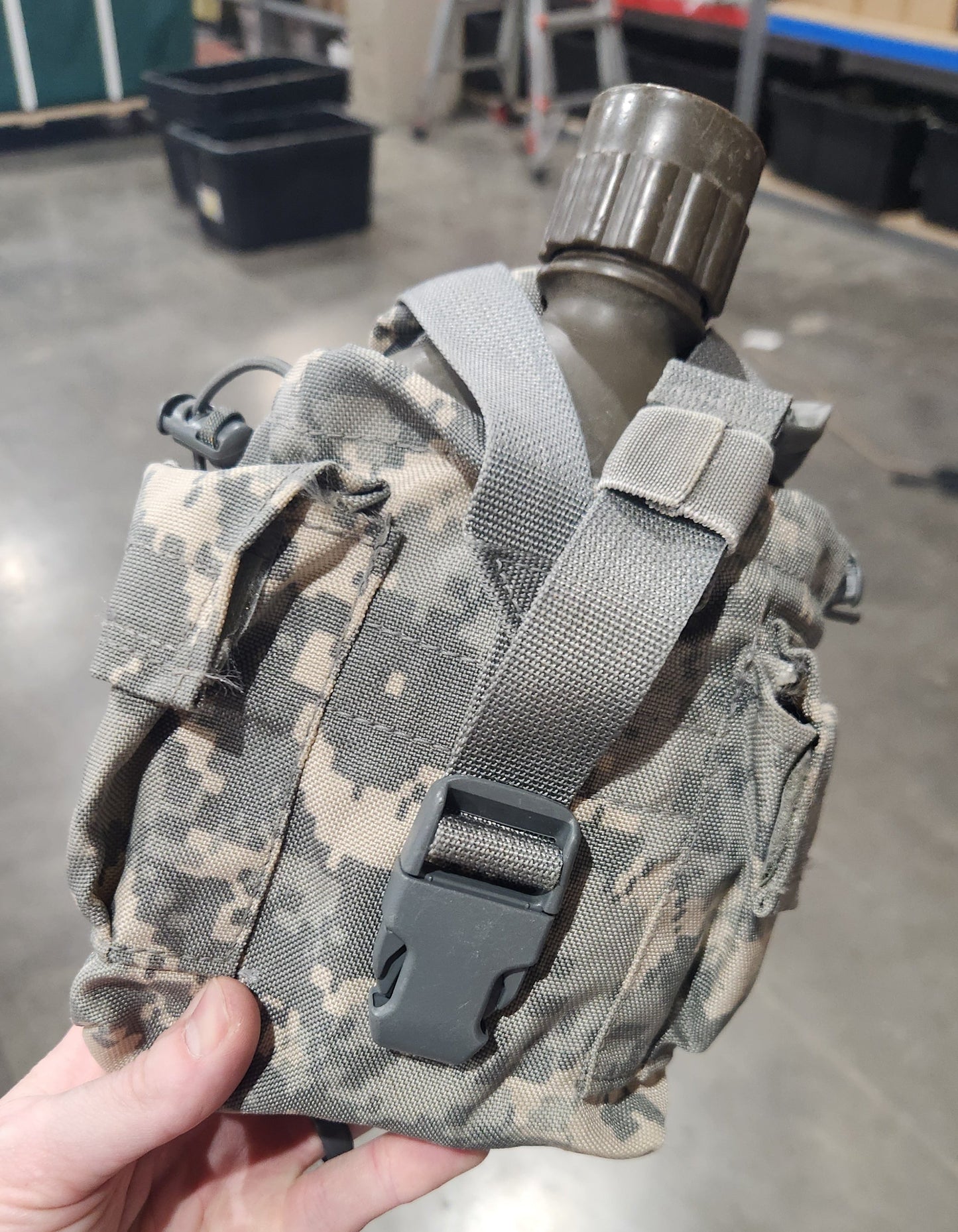 A hand holding a digital camouflage canteen pouch with a gray strap and a dark gray canteen bottle, set against a concrete warehouse floor.
