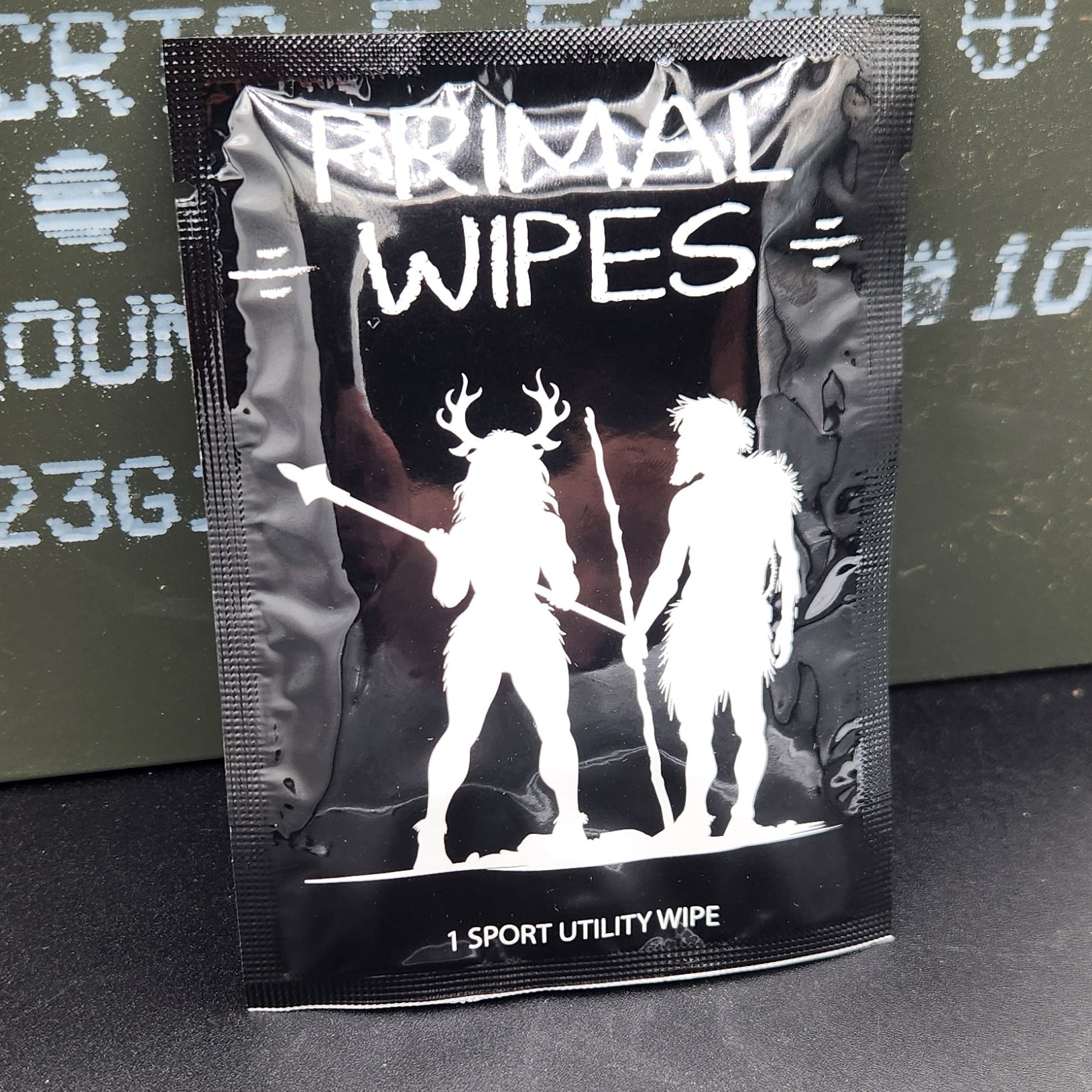 A small black packet labeled 'COLD LUNCH WIPES' with white graphics, placed on top of an olive green military-style container on a dark gray surface.