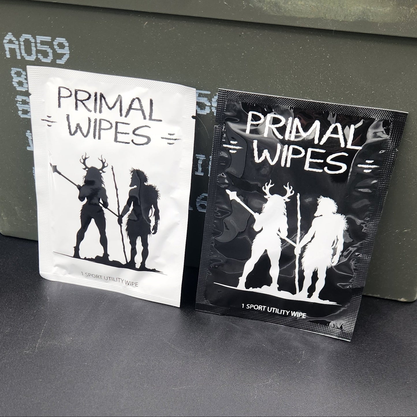 Two packs of Primal Wipes hand wipes, one black and one white, displayed on top of an olive green metal storage box against a dark gray background.