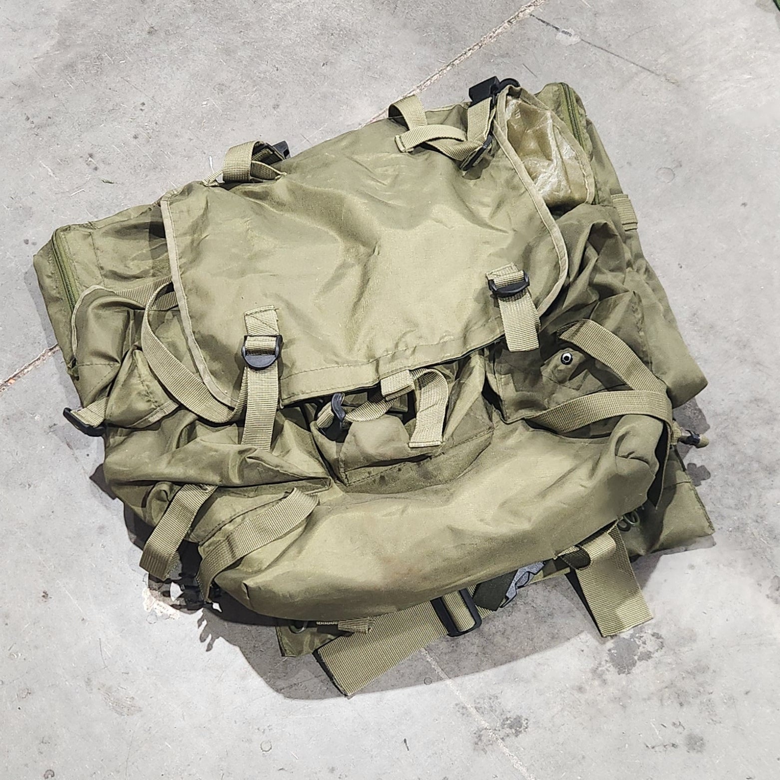 Green military-style backpack on a concrete floor with a person's foot visible.