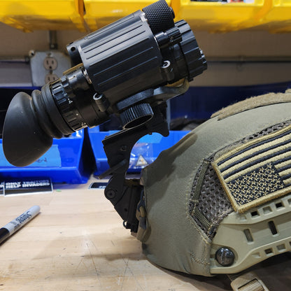 A black thermal or night-vision style scope with a large eyepiece and adjustment knobs, photographed attached to a tan tactical vest or backpack in a workshop or garage setting.