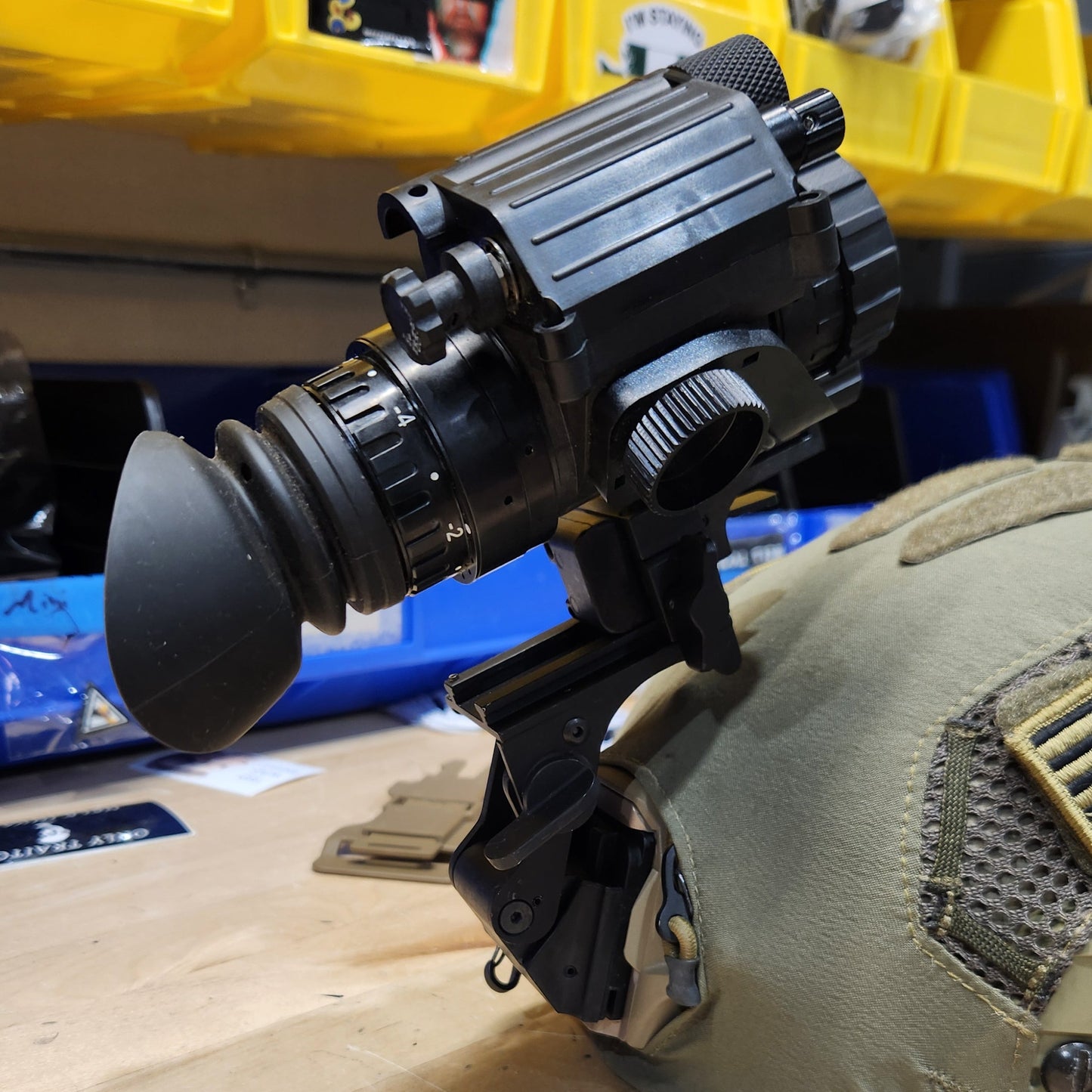 A black thermal or night vision scope mounted on a tan tactical vest, photographed in a workshop or storage area with yellow bins and a wooden wall visible.