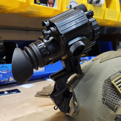 A black thermal or night vision scope mounted on a tan tactical vest, photographed in a workshop or storage area with yellow bins and a wooden wall visible.