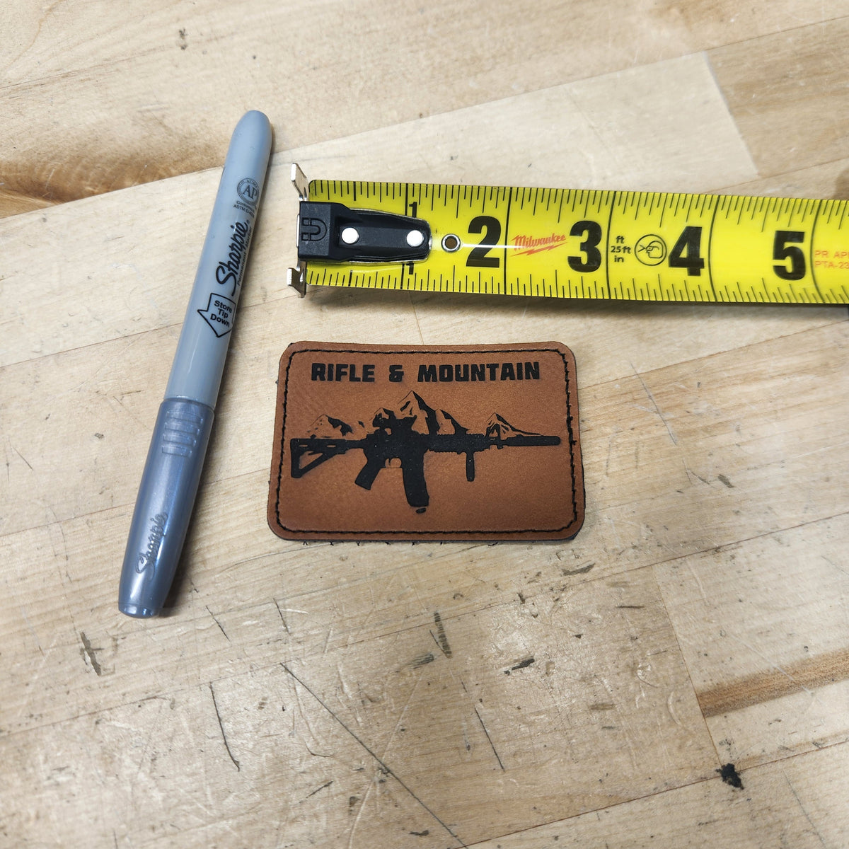 A brown rectangular leather patch with a black rifle graphic and the words 'RIFLE & MOUNTAIN' is shown on a light wood surface next to a gray Sharpie marker and a yellow tape measure.