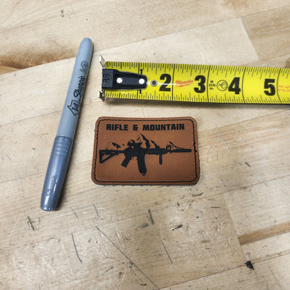 A brown rectangular leather patch with a black rifle graphic and the words 'RIFLE & MOUNTAIN' is shown on a light wood surface next to a gray Sharpie marker and a yellow tape measure.
