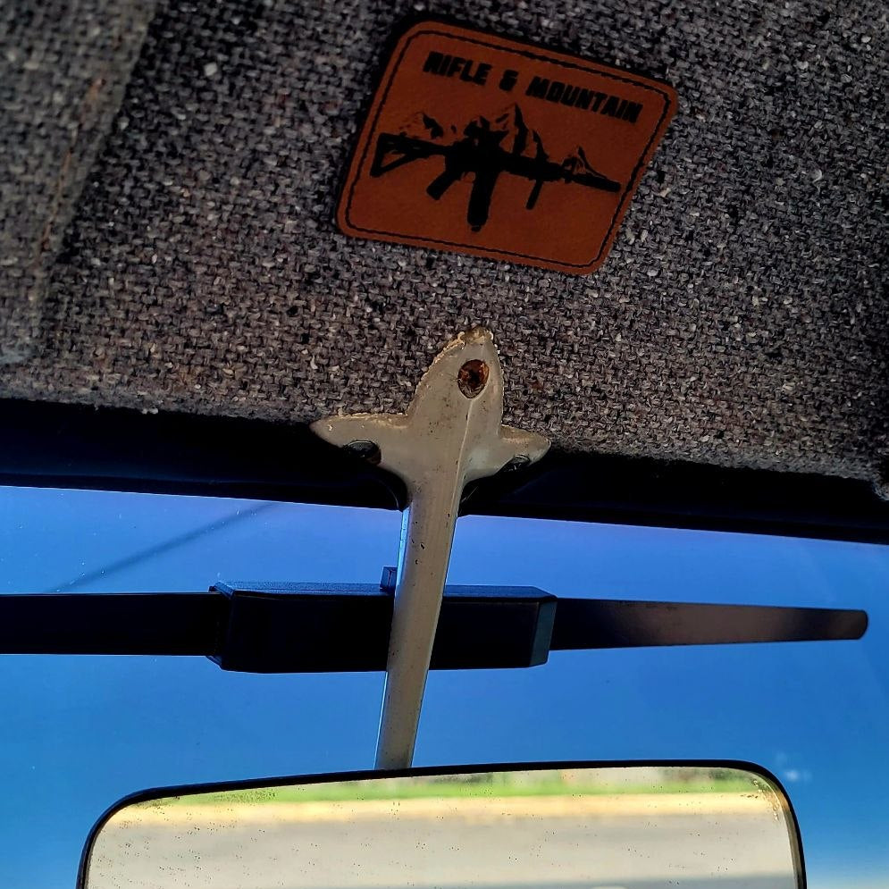 A close-up of a car interior showing a rearview mirror, windshield wiper arm, textured gray sunshade, and an orange-brown patch with a black rifle silhouette and the words 'RIFLE & MOUNTAIN'.
