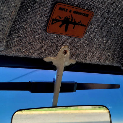 A close-up of a car interior showing a rearview mirror, windshield wiper arm, textured gray sunshade, and an orange-brown patch with a black rifle silhouette and the words 'RIFLE & MOUNTAIN'.