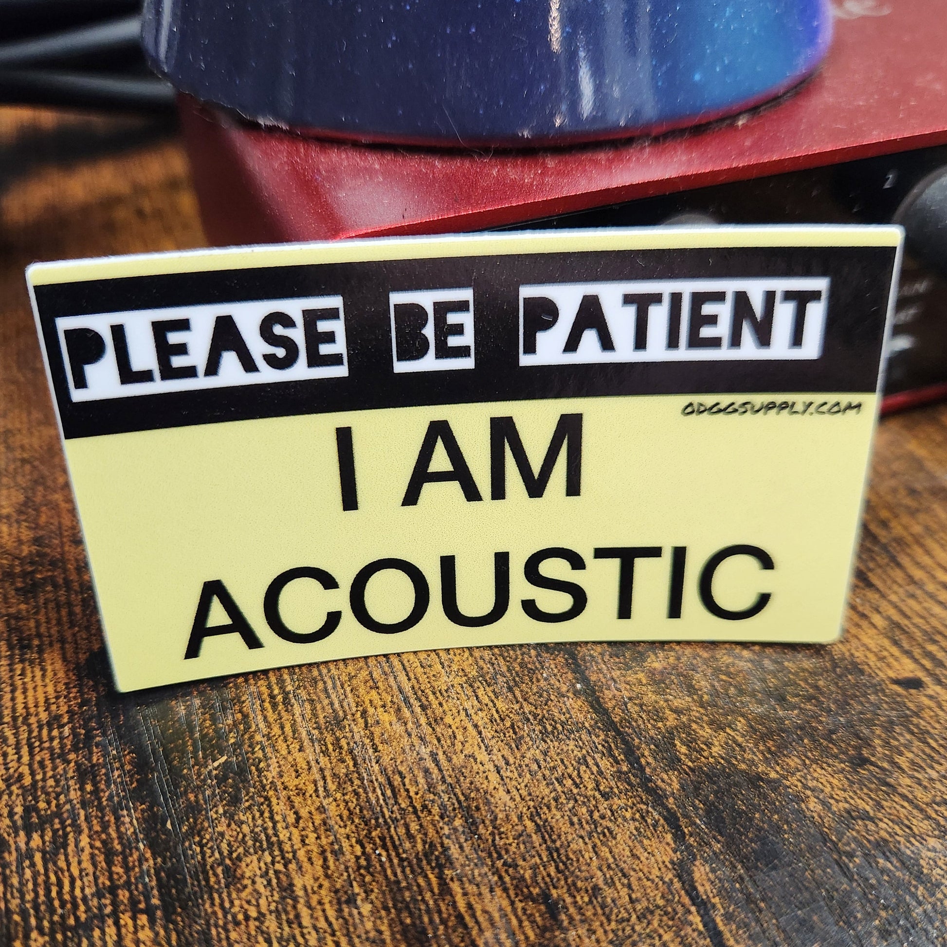 Sticker on a guitar neck with text 'Please be patient, I am acoustic' on a wooden surface.